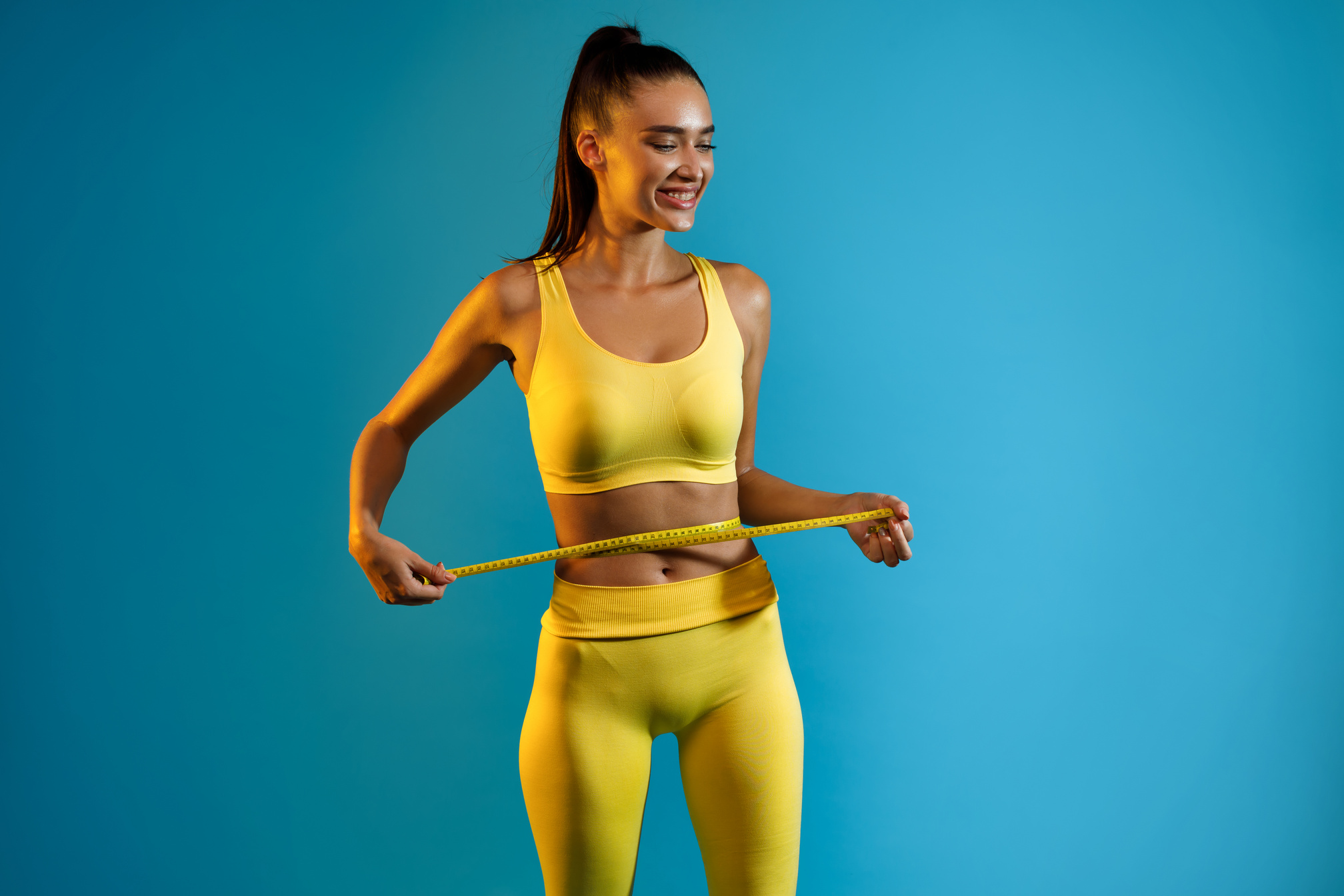 Fit Woman Measuring Waist with Tape after Weight-Loss, Blue Background